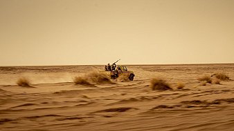 L'image montre un véhicule tout-terrain, probablement un pick-up, qui avance rapidement à travers un désert. La poussière s'élève autour de lui, créant une ambiance de mouvement et de vitesse. À l'arrière du véhicule, on aperçoit des silhouettes de personnes, probablement des passagers, qui semblent être équipés pour un environnement hostile. Le paysage est vaste et aride, avec des dunes de sable et des buissons résistant aux conditions sèches. Le ciel est clair mais teinté d'une couleur jaunâtre, suggérant une chaleur intense. L'ensemble donne une impression de solitude et d'aventure dans un milieu difficile.