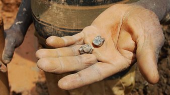 L'image montre une main, visiblement usée et sale, qui tient deux pierres. La paume est ouverte, révélant les deux morceaux de minéraux. L'un semble être plus gros et terne, tandis que l'autre est plus petit et a une surface légèrement brillante. Le fond de l'image montre un sol de terre battue, ce qui suggère que la scène se déroule dans un environnement minier ou rural. La lumière éclaire les pierres, mettant en avant leurs textures rugueuses et leurs formes irrégulières.
