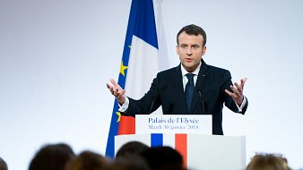 L'image montre un homme debout devant un podium, probablement en train de donner un discours. Il porte un costume sombre avec une cravate, et ses mains sont levées, ce qui indique qu'il met l'accent sur ses propos. En arrière-plan, on aperçoit le drapeau français aux couleurs bleu, blanc et rouge, ainsi qu'une autre bannière européenne. La scène semble se dérouler dans un cadre officiel, peut-être à l'Élysée, avec un public flou et attentif. L'atmosphère est sérieuse et engageante, marqué par le dynamisme du orateur.