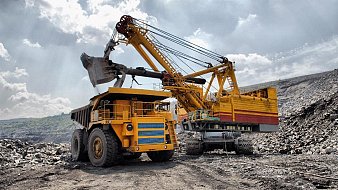 L'image présente un site d'extraction minière. Au premier plan, un gros camion de transport de couleur jaune, avec des roues massives, est chargé de matériaux. À côté, une grande pelle mécanique jaune et rouge, dotée d'un bras articulé, a relevé une charge de terre ou de roches. Le paysage environnant est accidenté, avec des tas de gravier et des pentes abruptes. Le ciel est partiellement nuageux, laissant passer des rayons de lumière qui illuminent la scène, créant une ambiance industrielle dynamique. Des bruits de machines et d'activités minières peuvent être imaginés, rendant l'environnement à la fois impressionnant et robuste.