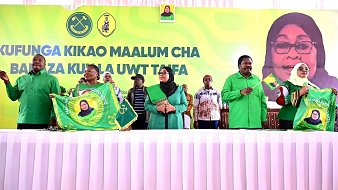 L'image montre un événement politique avec plusieurs personnes debout derrière une table, vêtues de chemises vertes. Au fond, on peut voir une grande bannière avec un portrait d'une femme souriante, probablement une dirigeante. Les participants tiennent des drapeaux ou des banderoles, souvent en vert et jaune, qui portent des symboles liés à leur organisation. L'atmosphère semble festive et engagée, avec des membres du public en arrière-plan qui écoutent attentivement. La réunion semble évoquer des thèmes de solidarité et de mobilisation.