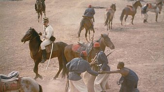 L'image présente une scène historique en plein air, où plusieurs personnages sont engagés dans une interaction dynamique. Au centre, un homme vêtu d'un uniforme militaire blanc est assis sur un cheval, observant la scène. Deux autres hommes, probablement des soldats, sont en train de se battre ou de grappler au sol, mettant en évidence un moment de tension. En arrière-plan, plusieurs autres chevaux et hommes se déplacent dans un environnement poussiéreux, suggérant une ambiance mouvementée et chaotique, typique d'un camp militaire. Le terrain est sec, peut-être de la terre battue, ce qui ajoute à l'atmosphère rustique et historique de la scène.