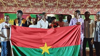 L'image présente un groupe de jeunes personnes rassemblées sous une tente décorée, avec des motifs colorés en arrière-plan. Au centre, ils tiennent une grande drapeau du Burkina Faso, qui est rouge avec une étoile verte au milieu. Les membres du groupe affichent des vêtements variés, certains portant des chemises traditionnelles, et semblent joyeux et solidaires. L'atmosphère est festive, suggérant un événement culturel ou une célébration.