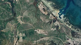 L'image représente une vue aérienne d'une région dont le paysage est marqué par des routes, des structures et des zones déforestées. On peut observer des chemins sinueux qui relient différents points, ainsi que des zones construites qui semblent être des installations industrielles ou militaires. La présence de la mer à droite de l'image ajoute un contraste avec la verdure terrestre, et on peut également voir des étendues d'eau et des zones côtières. Les nuances de couleur vont du vert des zones boisées aux teintes plus claires des infrastructures.