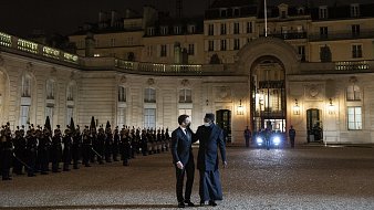 L'image représente une scène nocturne au Palais de l'Élysée, en France. Au premier plan, deux hommes discutent, l'un portant un costume noir et l'autre vêtu d'un long manteau sombre. En arrière-plan, une rangée de soldats en uniforme se tient en position, formant une haie d'honneur. Les bâtiments classiquement architecturés du palais sont illuminés, mettant en valeur leurs détails. Une lumière provenant d'un véhicule est visible à droite, ajoutant à l'atmosphère solennelle du moment. Les drapeaux français flottent au-dessus du bâtiment. L'ensemble de la scène dégage une impression de formalité et de dignité.