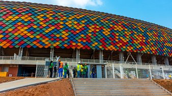 L'image montre un grand stade avec une architecture moderne. La façade du stade est ornée de tuiles de diverses couleurs vives, allant du rouge, orange, jaune, vert, bleu jusqu'au violet, formant une belle mosaïque sur un fond en béton. Au premier plan, on peut voir un groupe de personnes, habillées en vêtements de travail, qui semblent discuter ou se préparer à entrer dans le stade. Les escaliers mènent à une entrée principale, et le ciel au-dessus est bleu avec quelques nuages épars, ajoutant une impression de clarté et d'énergie à la scène.