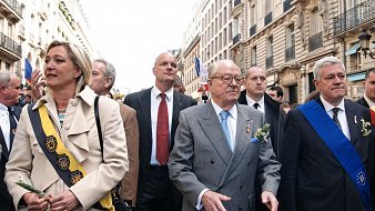 L'image montre un groupe de personnes marchant dans une rue animée, probablement lors d'un événement ou d'une cérémonie. Au centre, un homme plus âgé porte un costume gris clair et des lunettes. Il semble être une figure de proue, entouré de plusieurs autres individus, dont certains portent des sashes ou des écharpes distinctives, suggérant qu'ils occupent des rôles importants. On remarque également des décorations florales, indiquant une atmosphère cérémonielle. En arrière-plan, on aperçoit des bâtiments typiques, et des drapeaux flottent, ce qui évoque un rassemblement public festif et patriotique.