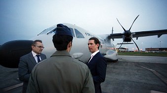 L'image représente un groupe de trois hommes se tenant près d'un avion sur une piste d'atterrissage. L'avion a des hélices et semble être un appareil de taille moyenne. L'éclairage est doux, indiquant qu'il peut être tôt le matin ou tard en soirée. Un homme en costume noir discute avec un autre homme habillé d'une tenue militaire. Le troisième homme, portant des lunettes, semble écouter la conversation. L'environnement est urbain et l'aéroport est visible en arrière-plan avec quelques lumières. L'atmosphère est professionnelle et engageante.