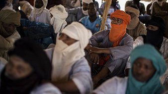 L'image montre un groupe de personnes assises dans un espace clos. Les individus portent des vêtements traditionnels, souvent en tissu léger, et sont partiellement masqués par des turbans ou d'autres pièces de tissu. On peut percevoir des couleurs variées : certains portent des turbans orange, d'autres bleus ou verts. Les visages des personnes sont en grande partie cachés, créant une atmosphère de mystère. L'environnement semble calme, et les positions des corps indiquent une attention collective. La lumière est douce, suggérant un éclairage naturel venant d'une source extérieure. On peut également sentir une ambiance de rassemblement, possiblement pour un échange culturel ou une discussion importante.