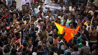 L'image représente une scène vibrante et animée d'une foule rassemblée ensemble. Au premier plan, on peut voir un groupe dense de personnes, principalement des jeunes hommes, exprimant leur énergie et leur enthousiasme. Certains d'entre eux lèvent les bras, d'autres applaudissent ou tiennent des drapeaux aux couleurs vives, notamment le rouge et le jaune. Au centre, une personne semble parler ou chanter, attirant l'attention de la foule. Les visages expriment des émotions variées telles que la joie, la passion et la détermination. Par temps peut-être nuageux, un parapluie coloré est en arrière-plan, ajoutant une note de couleur à la scène. L'ambiance générale est celle d'un rassemblement convivial, où les participants partagent un moment fort ensemble.
