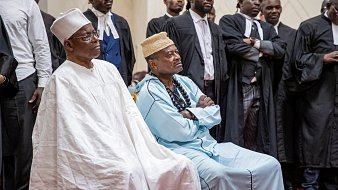 L'image montre un groupe de personnes rassemblées dans un espace formel, probablement un tribunal ou un événement solennel. Au premier plan, deux hommes sont assis. L'un porte une tenue traditionnelle blanche avec un chapeau, tandis que l'autre est habillé d'une tunique bleue et d'un collier de perles. Autour d'eux se trouvent des hommes en costume noir, certains portant des robes d'avocat. Tous semblent attentifs et sérieux, ce qui suggère que l'événement en cours est d'une grande importance. L'ambiance générale peut être perçue comme solennelle, avec une concentration visible sur les visages des participants.