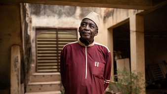 L'image montre un homme âgé portant une tenue traditionnelle d'une couleur rouge foncé. Sa chemise longue est agrémentée de détails blancs. Il se tient debout, les bras le long du corps, et regarde légèrement vers le haut. L'arrière-plan présente un bâtiment avec des murs usés et une porte en bois, ainsi que quelques plantes verdoyantes qui ajoutent une touche de nature à la scène. L'homme dégage une expression calme et réfléchie, créant une atmosphère de sagesse et de sérénité.