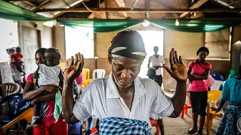 L'image montre un intérieur de salle, où des chaises colorées sont disposées de manière informelle. Au premier plan, une femme se tient debout, les bras levés, dans une posture de prière ou de méditation. Elle porte un foulard sur la tête et une blouse blanche, tandis qu'une jupe à motifs colorés entoure sa taille. En arrière-plan, on aperçoit d'autres personnes, certaines sont engagées dans une conversation tandis que d'autres semblent également en prière. L'ambiance est empreinte de spiritualité et d'un sentiment de communauté. La lumière naturelle entre par des fenêtres, créant un espace à la fois chaleureux et intime.