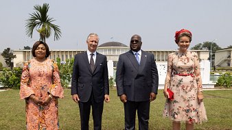 L'image montre un groupe de quatre personnes se tenant debout dans un jardin bien entretenu. À gauche, une femme porte une robe longue à motifs floraux et des manches évasées. À côté d'elle, un homme en costume sombre est debout, affichant une expression chaleureuse. Ensuite, un autre homme, également en costume, a une posture amicale et est au centre de l'image. Finalement, à droite, une femme porte une robe à fleurs avec un joli chapeau assorti et un sac à main rouge, se tenant avec élégance. En arrière-plan, on aperçoit un bâtiment imposant et un palmier qui ajoutent une atmosphère exotique à la scène. Le ciel est clair, ce qui contribue à un sentiment de rencontre officielle.