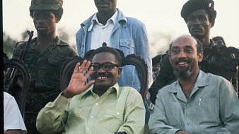 L'image présente un groupe de personnes, dont deux hommes au premier plan, assis sur des chaises ornées. L'un des hommes, souriant et portant des lunettes, salue de la main. Il a une chemise verte claire. À sa droite, un autre homme avec une chemise beige sourit également. En arrière-plan, plusieurs soldats en uniforme de camouflage et avec des casquettes se tiennent debout. L'environnement semble être en plein air, peut-être lors d'un événement officiel, avec un ciel illuminé par le crépuscule.