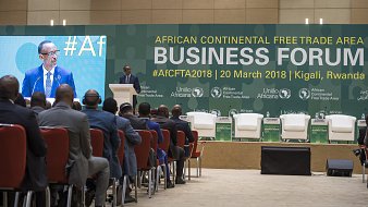 L'image présente un forum d'affaires qui se tient dans le cadre de la Zone de libre-échange continentale africaine. Au premier plan, on voit un homme, vraisemblablement un orateur, debout au micro, s'adressant au public. Il est entouré d'une scène bien agencée avec plusieurs chaises blanches et des logos des organisations impliquées dans l'événement. Le public, de dos, est composé d'hommes en costume, attentifs à la présentation. L'ambiance semble formelle et axée sur les discussions économiques. En arrière-plan, des banderoles mentionnent le forum et sa date, soulignant l'importance de l'événement pour le commerce en Afrique.