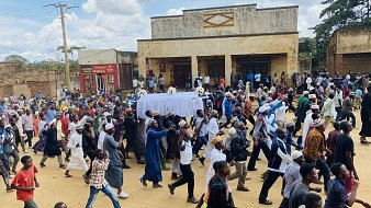 L'image montre une scène animée dans une rue où une grande foule se déplace. Au centre de l'attention, des personnes portent un drap blanc qui semble recouvrir un cercueil, indiquant qu'il s'agit d'un cortège funéraire. Les membres de la foule sont vêtus de divers habits, certains en tenues traditionnelles, d'autres plus contemporaines. Les visages reflètent des émotions variées, allant du recueillement à la tristesse. À l'arrière-plan, on peut remarquer des bâtiments aux murs fissurés, avec des panneaux publicitaires visibles. Le ciel est légèrement nuageux, créant une atmosphère à la fois sombre et lugubre, mais la scène reste vibrante de vie grâce à la présence de nombreux participants.