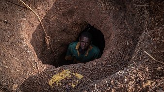 Dans cette image, nous voyons un homme qui se tient au fond d'un trou creusé dans le sol. Le trou a une forme ronde, et les parois sont en terre brune, légèrement humides et irrégulières. L'homme, vêtu d'un maillot vert avec un col jaune, regarde vers le haut avec une expression sérieuse. Son visage est en partie dans l'ombre, mais son regard est visible. Autour du trou, on peut apercevoir des gravats et des brindilles qui ajoutent un aspect naturel et brut à l'environnement. La scène évoque un sentiment d'isolement et de travail ardu.