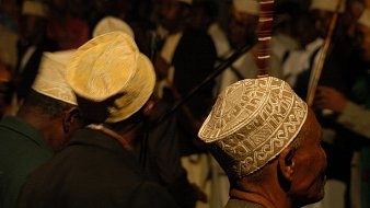 L'image présente un groupe de personnes, principalement des hommes, de profil. Ils portent des chapeaux traditionnels, souvent ornés de motifs délicats, qui varient en couleurs et en designs. En arrière-plan, on peut distinguer d'autres figures et instruments de musique, suggérant une ambiance festive ou rituelle. La lumière est tamisée, créant une atmosphère intime. Les visages des hommes sont flous, mais leur posture et leurs chapeaux ajoutent une touche culturelle à la scène.