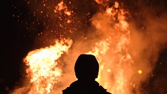 L'image montre une scène nocturne où une personne se tient de dos, face à un grand feu. Les flammes dansent avec éclat, projetant une lumière vive et chaleureuse dans l'obscurité environnante. Des étincelles s'élèvent en spirales, créant une ambiance à la fois magique et intense. La silhouette de la personne est sombre, contrastant avec les couleurs vives de l'incendie, ce qui permet de ressentir la chaleur émanant du feu et l'atmosphère captivante qui l'entoure.