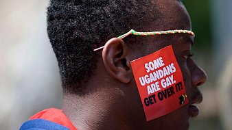 L'image représente un homme dont le visage est de profil. Il porte un petit autocollant sur sa joue qui affiche un message en lettres blanches sur fond rouge. Le texte dit : "SOME UGANDEANS ARE GAY. GET OVER IT!" (Certains Ougandais sont gays. Acceptez-le !). L'homme a des cheveux courts et un accessoire coloré autour de la tête. L'ambiance générale de l'image suggère un acte de protestation ou de revendication des droits.