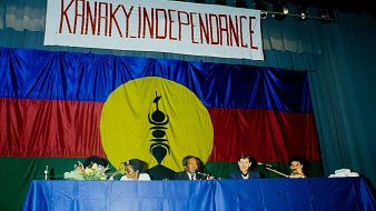 L'image présente une scène de réunion ou de conférence, où des personnes sont assises derrière une table. En arrière-plan, il y a un grand drapeau avec des couleurs vives : un rouge et un bleu, et un cercle jaune au centre. Dans ce cercle, on peut distinguer un symbole noir, précisant ainsi l'identité culturelle. Au-dessus de la scène, une banderole arbore le mot "KANAKY INDEPENDANCE" écrit en lettres rouges, exprimant un message fort lié à l'indépendance. Les participants semblent engagés et sont en discussion, créant une atmosphère de détermination et de solidarité.