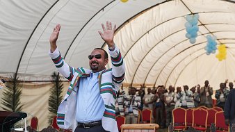 L'image montre un homme, probablement un leader, se tenant debout sous un grand chapiteau. Il porte une tenue colorée avec des rayures multicolores et des lunettes de soleil, et lève les deux mains dans un geste de salutation ou d'encouragement. En arrière-plan, on aperçoit un groupe de personnes qui semblent l'écouter attentivement, tous habillés de manière décontractée. Le chapiteau est décoré avec des ballons, ajoutant une ambiance festive à l'événement. L'atmosphère générale est celle d'un rassemblement joyeux et engageant.