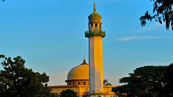 L'image montre un groupe de personnes en train de prier ensemble. Au centre, un homme en costume bleu se tient les mains jointes, la tête légèrement inclinée, montrant une attitude de recueillement. Autour de lui, plusieurs hommes portent des vêtements traditionnels, incluant des turbans et des chapeaux décorés. Tous semblent concentrés et tournés vers l'avant, les yeux fermés ou la tête baissée, dans un moment de méditation ou de prière. L'ambiance est empreinte de sérénité et de respect.