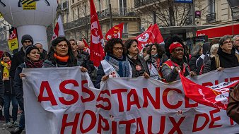 L'image montre une manifestation dans une rue animée. Au premier plan, un groupe de personnes tient une grande banderole blanche sur laquelle est écrit "ASSISTANCE HÔPITAUX DE…" en lettres rouges. Les manifestants arborent des sourires et semblent déterminés. On aperçoit également plusieurs drapeaux rouges, qui sont souvent associés à des syndicats. L'atmosphère est dynamique, avec une multitude de personnes autour, représentant la solidarité et l'engagement. Les bâtiments en arrière-plan suggèrent une ambiance urbaine, typique des grandes villes.