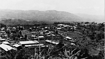 Cette image propose une vue panoramique d'un paysage rural. Au premier plan, on remarque une végétation dense, avec des feuilles larges et des arbres. En arrière-plan, se déploie une vallée avec plusieurs maisons aux toits en tôle, éparpillées sur le terrain. Certaines constructions sont entourées d'arbres et de collines, tandis que les montagnes s'élèvent au loin, créant un contraste avec le ciel. Les nuances de gris de l'image évoquent un atmosphère nostalgique et calme, typique d'une région tranquille, loin du brouhaha urbain.