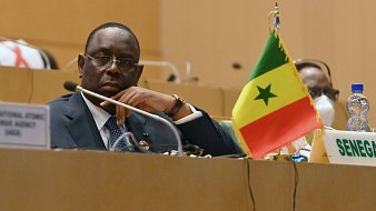 L'image montre un homme assis dans un cadre officiel, probablement lors d'une réunion ou d'une conférence. Il porte un costume sombre avec une chemise et une cravate, et affiche une expression pensive ou attentive. À sa droite, un petit drapeau du Sénégal est visible, avec les couleurs rouge, jaune et vert, ainsi que l'étoile verte au centre. On peut également apercevoir une bouteille d'eau à gauche et un badge ou un panneau indiquant "SENEGAL" devant lui. L'ambiance générale est sérieuse et concentrée, typique de discussions diplomatiques ou politiques.