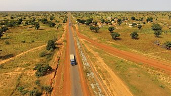 Cette image présente un paysage rural étendu, où une route goudronnée traverse une vaste plaine. Sur la droite, on aperçoit un camion bleu qui circule, indiquant une activité de transport. De part et d'autre de la route, le sol est d'un rouge terreux, typique de certaines régions africaines, semé de petites collines et de végétation éparse, notamment des arbres et des buissons. Sur la gauche de l'image, on peut distinguer des habitations simples, suggérant la présence d'un village. L'arrière-plan est composé de champs et d'arbres, renforçant l'impression de tranquillité et d'espace ouvert. L'ensemble évoque une atmosphère de ruralité, avec une nature généreuse, baignée par une lumière intense.