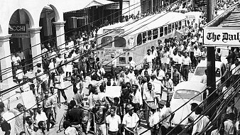 L'image que vous avez devant vous représente une scène urbaine animée dans les années 1960. On peut voir une foule dense de personnes marchant dans une rue. Les gens portent des vêtements typiques de l'époque, et il semble qu'ils se déplacent avec un but précis. À gauche, plusieurs commerces sont visibles, avec des enseignes en lettres visibles. Un ancien bus à l'allure caractéristique est garé, ajoutant une touche nostalgique à la scène. On aperçoit également quelques voitures stationnées et d'autres qui circulent dans la rue. Les fils électriques sont visibles en haut de l'image, accentuant l'impression d'une ville vivante et dynamique. L'atmosphère générale évoque une période de mouvement socioculturel important, avec des échanges et une vitalité palpable dans l'air.