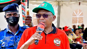 L'image montre un homme qui prend la parole devant un public. Il porte un t-shirt rouge avec le texte "ANNIVERSAIRE DU PARTI" et une casquette verte. Il tient un micro et semble s'adresser à l'assemblée avec passion. À l'arrière-plan, on peut apercevoir des gens assis, certains portant des vêtements colorés. Un agent de sécurité en uniforme bleu est visible à côté de lui, ajoutant un élément de protection ou d'autorité à l'événement. L'atmosphère semble festive et engagée.