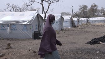 L'image montre un camp de réfugiés, caractérisé par plusieurs tentes en toile blanches et bleues, portant le logo de l'UNHCR. Au premier plan, une femme marche, vêtue d'un voile mauve et d'une robe claire, se dirigeant vers l'une des tentes. Le paysage environnant est aride et désolé, avec quelques arbres dénudés, suggérant un environnement difficile. En arrière-plan, des lignes électriques sont visibles, ce qui indique une certaine infrastructure. L'atmosphère semble calme, mais il y a une sensation de précarité et de résilience.
