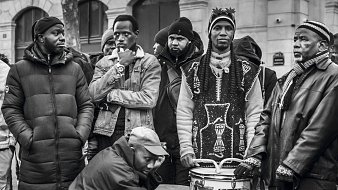 L'image montre un groupe de plusieurs hommes rassemblés dans un environnement urbain. Ils portent des vêtements variés, allant de parkas sombres à des vêtements traditionnels. Certains d'entre eux regardent dans des directions différentes, affichant une diversité d'expressions. Au centre, un homme est assis, jouant sur un tambour, ajoutant une ambiance musicale à la scène. Les autres semblent attentifs, créant une atmosphère de camaraderie et d'échange. L'image est en noir et blanc, accentuant les contrastes et les textures des vêtements et des visages.