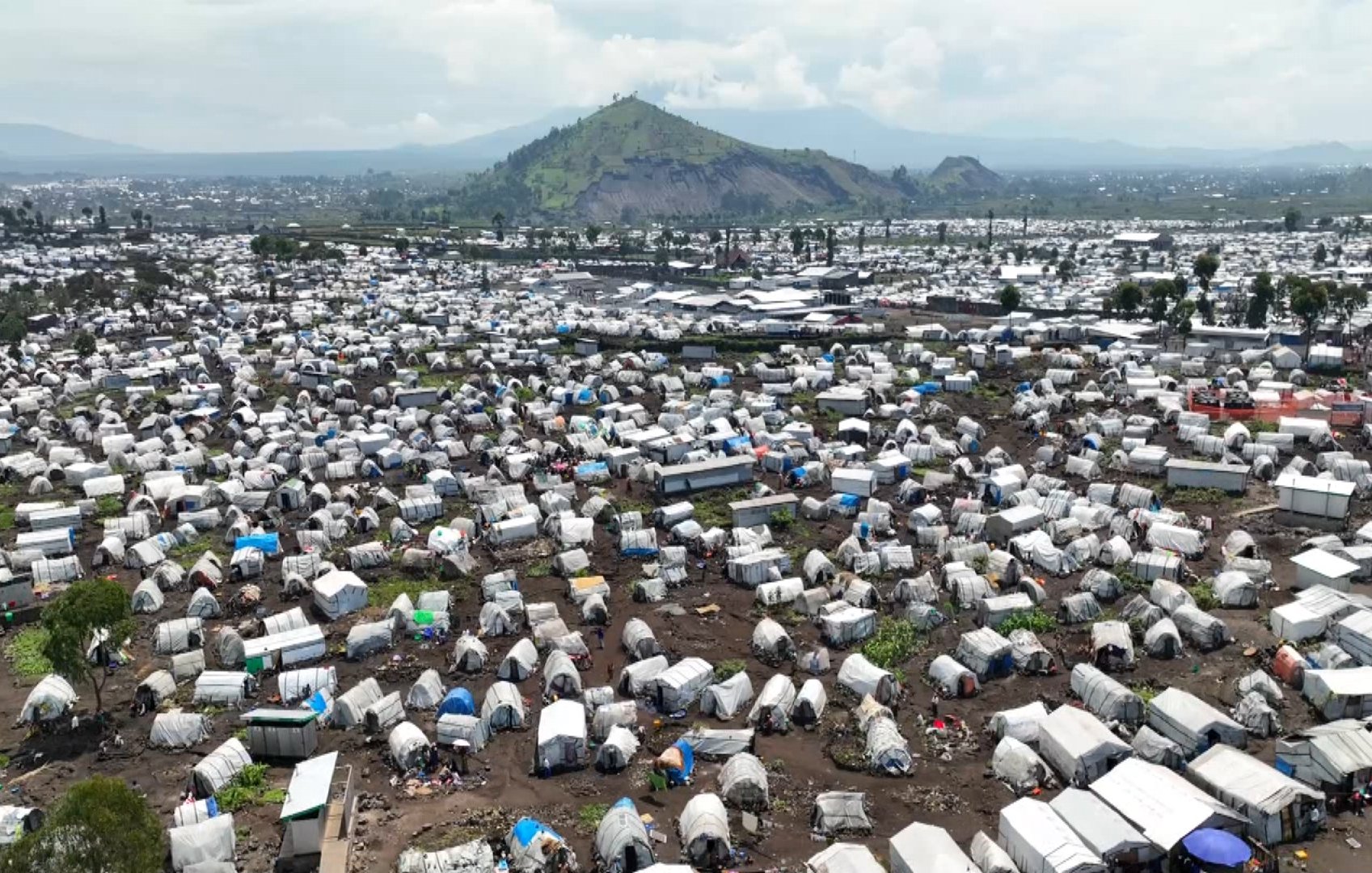 L'image montre un vaste camp de tentes, probablement un site de secours ou un camp de réfugiés, s'étendant à perte de vue. Les tentes sont de couleurs multiples, avec des toits en toile qui varient du blanc au bleu, et certaines sont en désordre avec des morceaux de tissu et de plastique. Le terrain est accidenté, avec des traces d'usage et de la terre visible entre les tentes. En arrière-plan, on aperçoit une montagne verdoyante qui domine le paysage, ajoutant un contraste entre la nature et l'afflux humain. À distance, on distingue des constructions urbaines, des routes et d'autres installations, suggérant la proximité d'une ville. L'ensemble de la scène véhicule une impression de densité et de survie collective, caractéristique des situations d'urgence humanitaire.