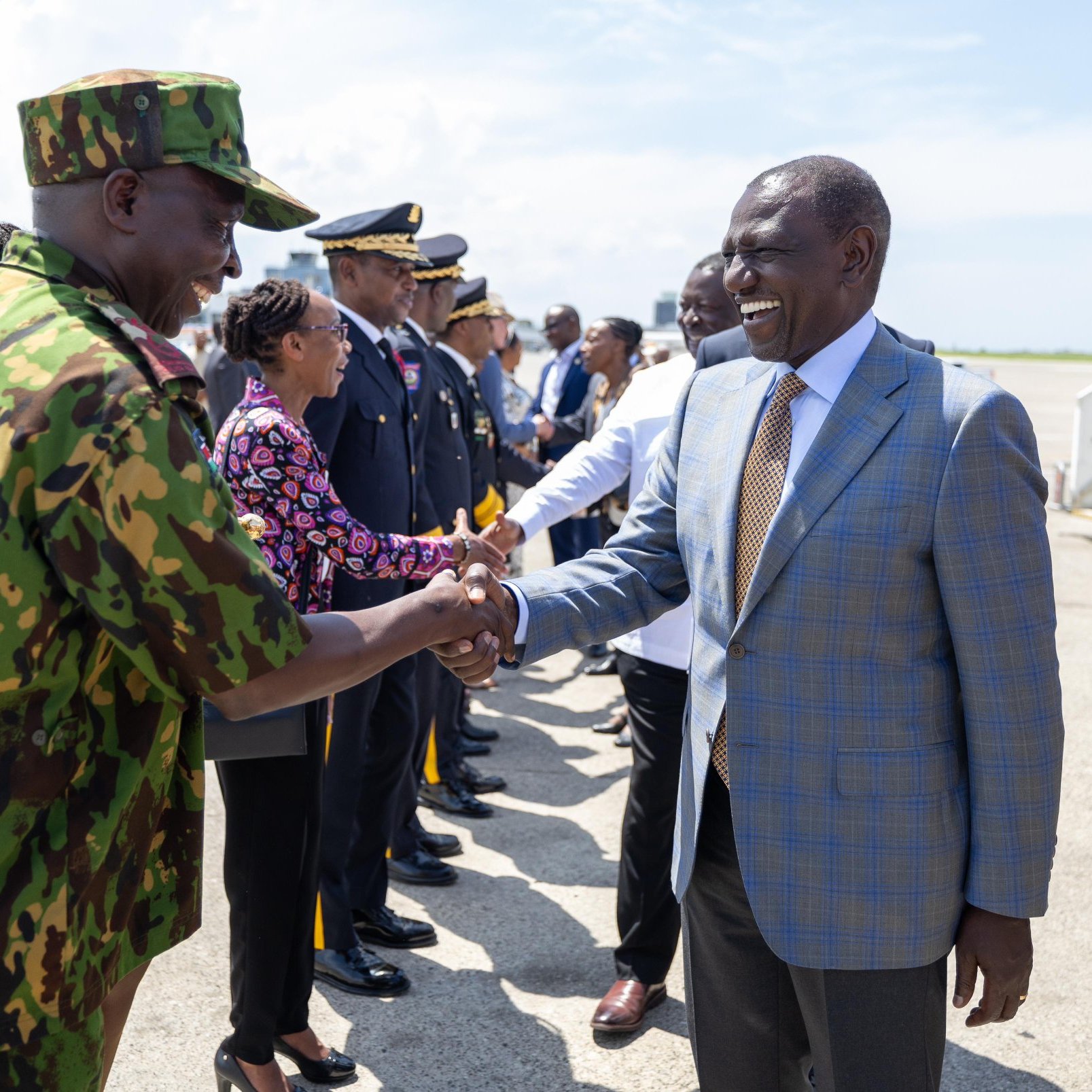 L'image montre une scène de bienvenue officielle à l'aéroport. Au premier plan, deux hommes se serrent la main, exprimant une atmosphère de camaraderie et de respect. L'un des hommes est en uniforme militaire avec un motif camouflage, tandis que l'autre porte un costume élégant avec une cravate. Autour d'eux, un groupe de personnalités, dont des membres des forces de sécurité et des fonctionnaires en civil, se tient en ligne, attendant leur tour pour saluer. En arrière-plan, le ciel est clair et ensoleillé, créant une ambiance positive. L'ensemble de la scène respire la solennité et l'importance d'un événement officiel.