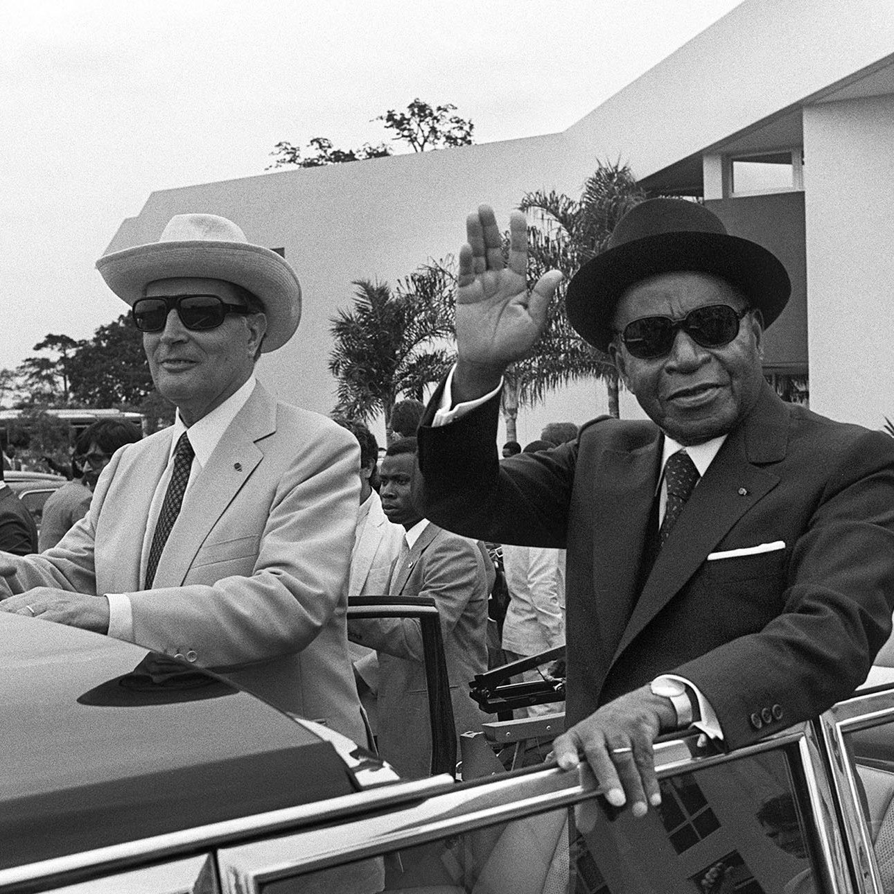 L'image représente deux hommes à l'extérieur d'une voiture décapotable. L'environnement semble être un événement officiel ou une cérémonie, avec des bâtiments modernes en arrière-plan. Le premier homme, à gauche, porte un costume clair, un chapeau de paille et des lunettes de soleil. Il a une expression sérieuse et regardent vers l'avant. Le second homme, à droite, est vêtu d'un costume foncé, d'un chapeau noir et également de lunettes de soleil. Il lève la main en signe de salut, avec un léger sourire. Il y a une foule de personnes en arrière-plan, habillées en costumes formels, ce qui indique une atmosphère festive ou cérémonielle. Des palmiers ou des arbres peuvent être aperçus, ajoutant une touche tropicale à l'environnement.