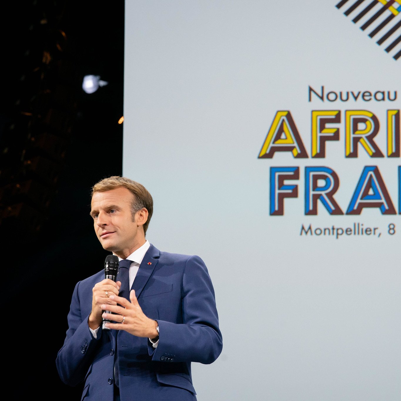 L'image montre un homme, probablement un leader ou un politicien, en train de s'adresser à un public. Il porte un costume sombre et une cravate, et il tient un micro dans sa main. En arrière-plan, il y a un grand écran avec le texte "Nouveau Sommet AFRIQUE FRANCE" suivi de la mention "Montpellier, 8 octobre 2021". L'ambiance est formelle et l'accent est mis sur la rencontre entre la France et les pays africains. Le public semble attentif à son discours.