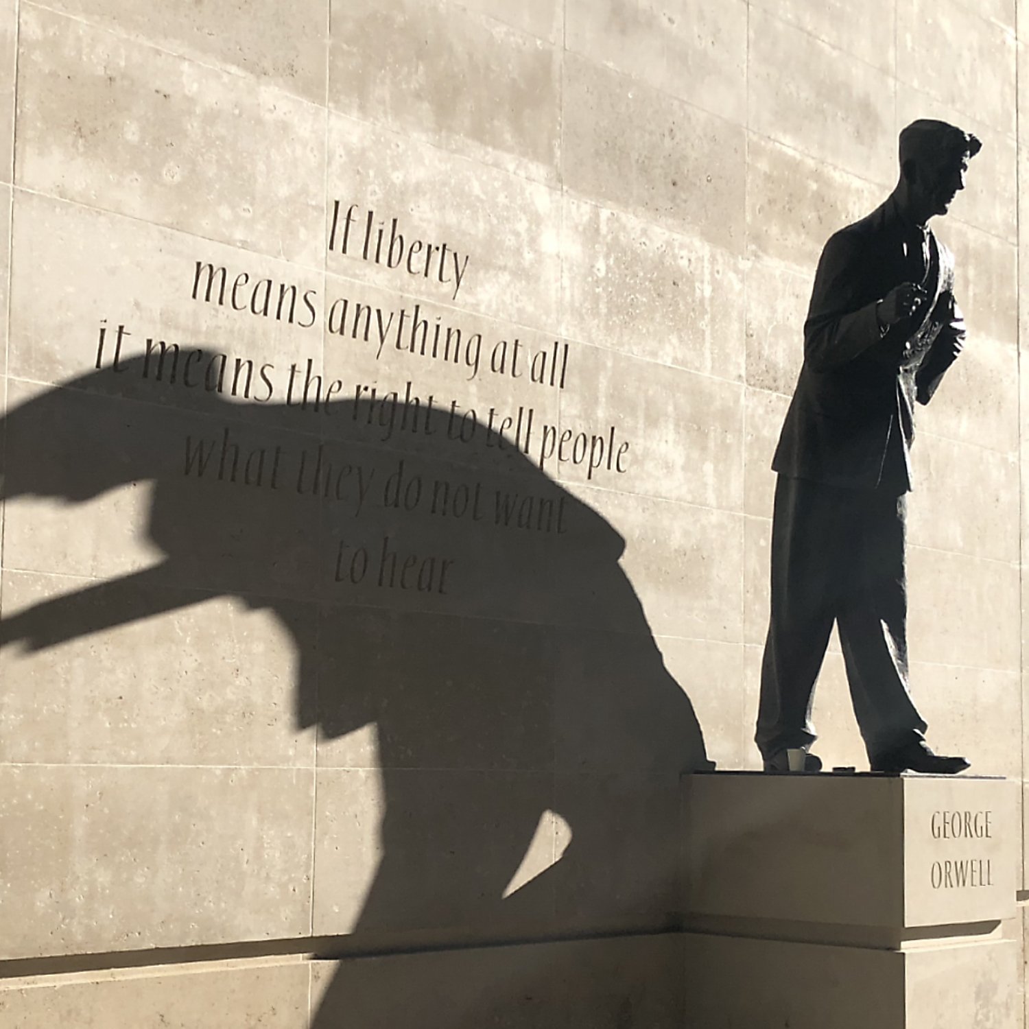 L'image représente une statue d'un homme debout, probablement un auteur ou un penseur, sur un socle. La statue est en bronze, et son ombre projette une forme dynamique sur le mur derrière elle. Ce mur est en pierre lisse, et il porte une citation célèbre qui commence par « If liberty means anything at all... », reflétant des thèmes de liberté et d'expression. L'ensemble crée une atmosphère réfléchie et inspirante, incitant à considérer la profondeur des mots inscrits ainsi que le rôle de la statue.