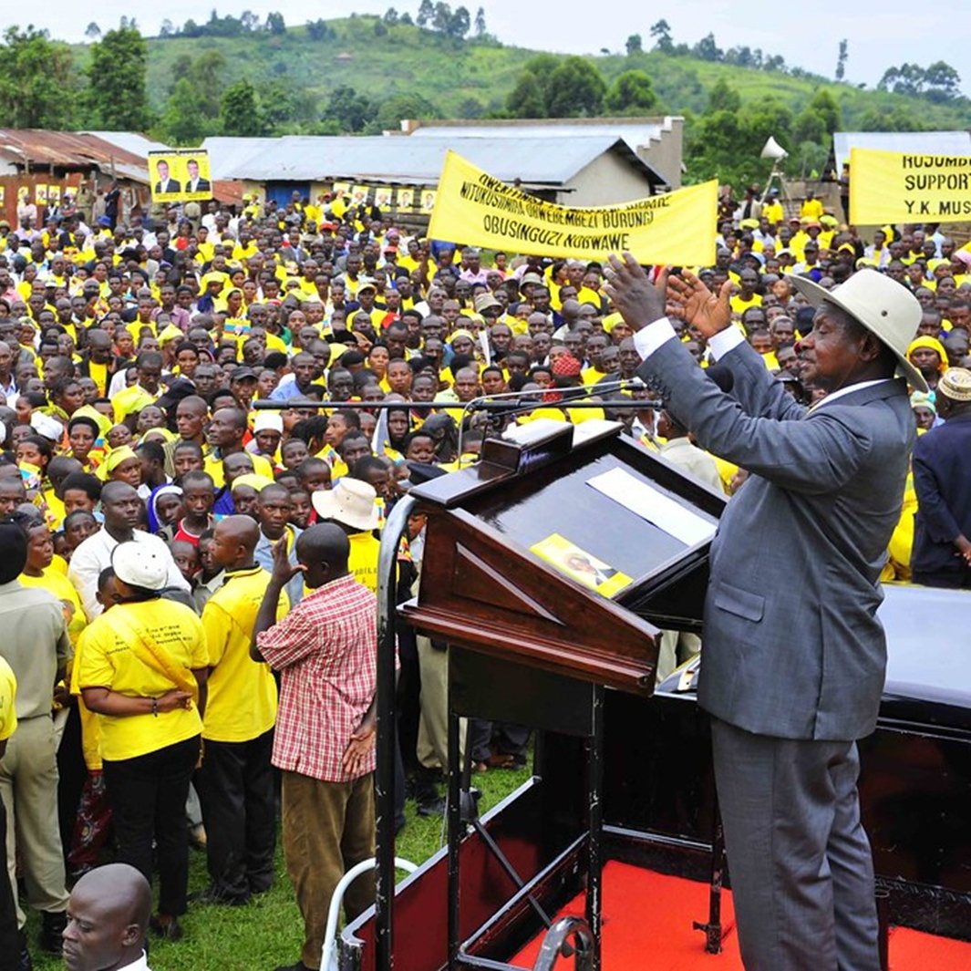 L'image montre une grande foule rassemblée en plein air. Les personnes dans la foule portent majoritairement des vêtements jaunes, ce qui pourrait indiquer qu'elles soutiennent un mouvement ou un parti spécifique. Au premier plan, un homme, vraisemblablement un orateur ou un dirigeant, se tient derrière un podium. Il lève les bras, semblant interagir avec la foule et exprimer un message puissant. En arrière-plan, on aperçoit des bâtiments simples et des collines verdoyantes, donnant un cadre naturel à l'événement. L'atmosphère semble énergique et mobilisée.