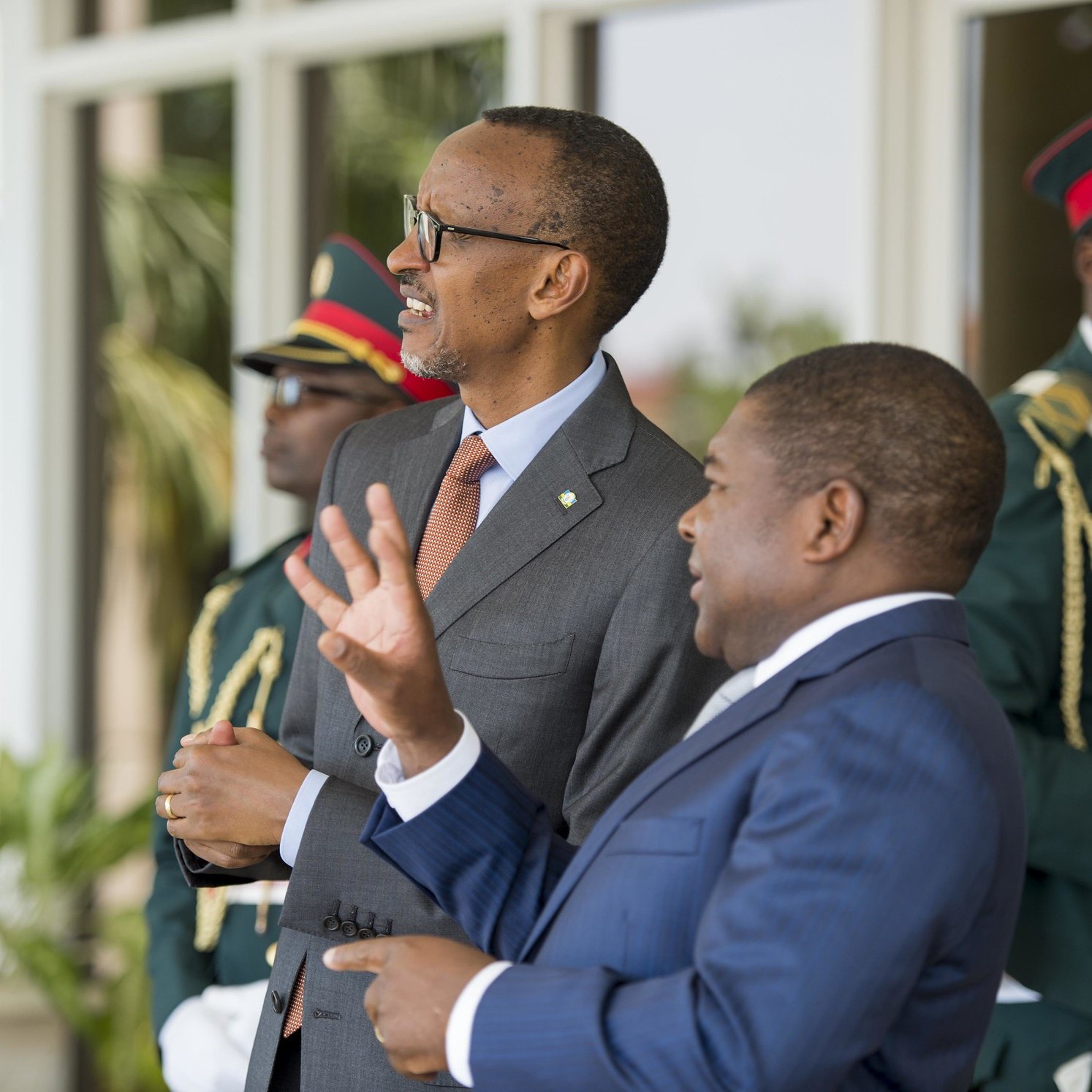 Sur cette image, nous voyons deux hommes en costume dialoguant dans un cadre officiel. L'homme à gauche, avec des lunettes et un sourire, semble être en train d'écouter attentivement. À sa droite, un homme aux cheveux courts gesticule avec les mains, soulignant un point dans la conversation. En arrière-plan, deux gardes en uniforme militaire se tiennent, assurant une ambiance de sécurité. On peut également percevoir des éléments de verdure, suggérant un environnement extérieur agréable.
