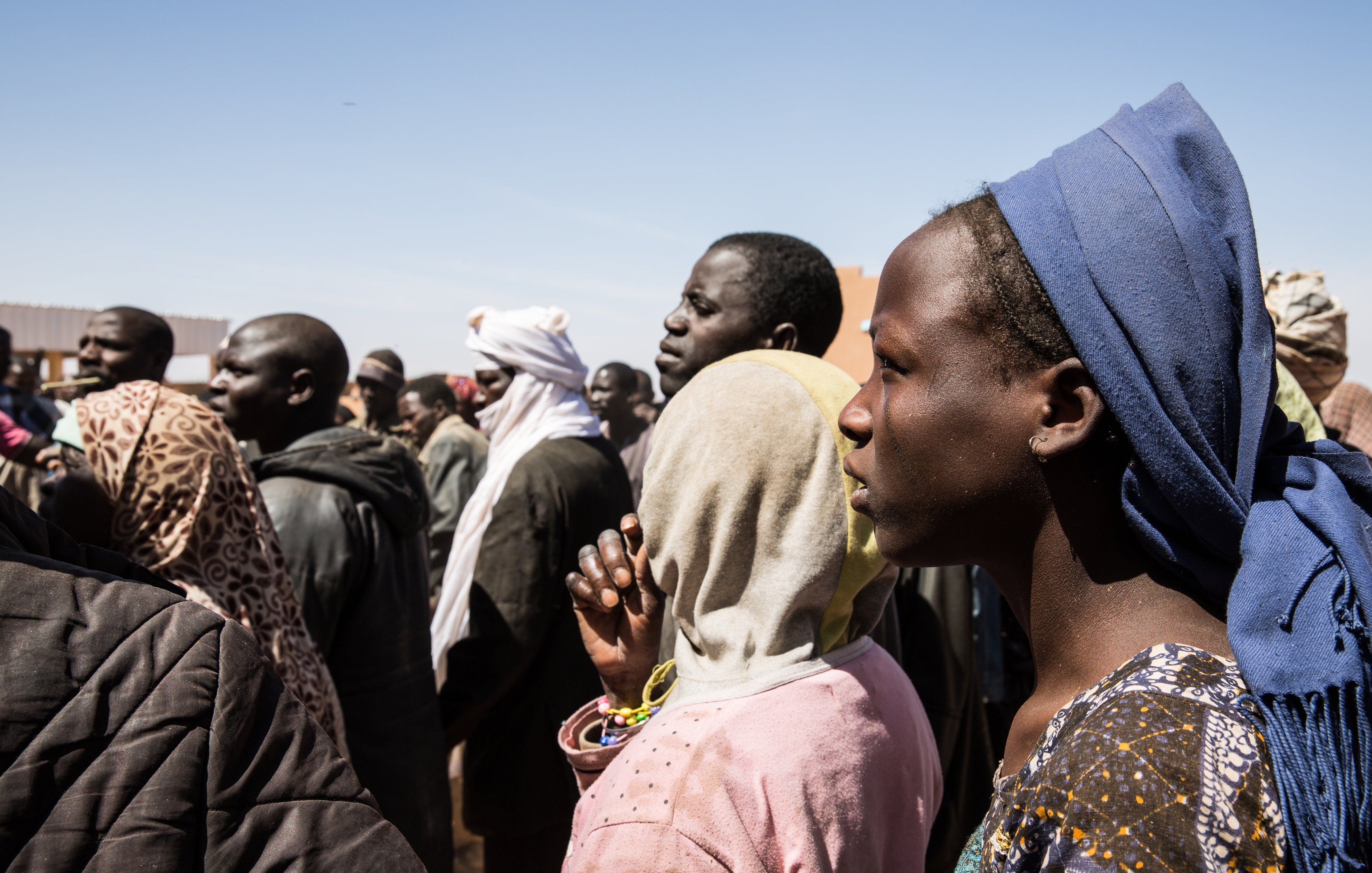 L'image montre un groupe de personnes se tenant debout, la plupart tournées vers un même point. On peut distinguer des hommes et des femmes de différents âges, habillés de vêtements variés, souvent colorés et traditionnels. Certaines personnes portent des couvertures sur la tête ou des scarves. L'expression sur leurs visages semble sérieuse, indiquant peut-être une attente ou une attention particulière. L'arrière-plan présente un ciel clair, suggérant une journée ensoleillée, et des structures émergent faiblement en arrière-plan, ajoutant à l'environnement d'un rassemblement. Le moment semble chargé d'émotions et de tension collective.