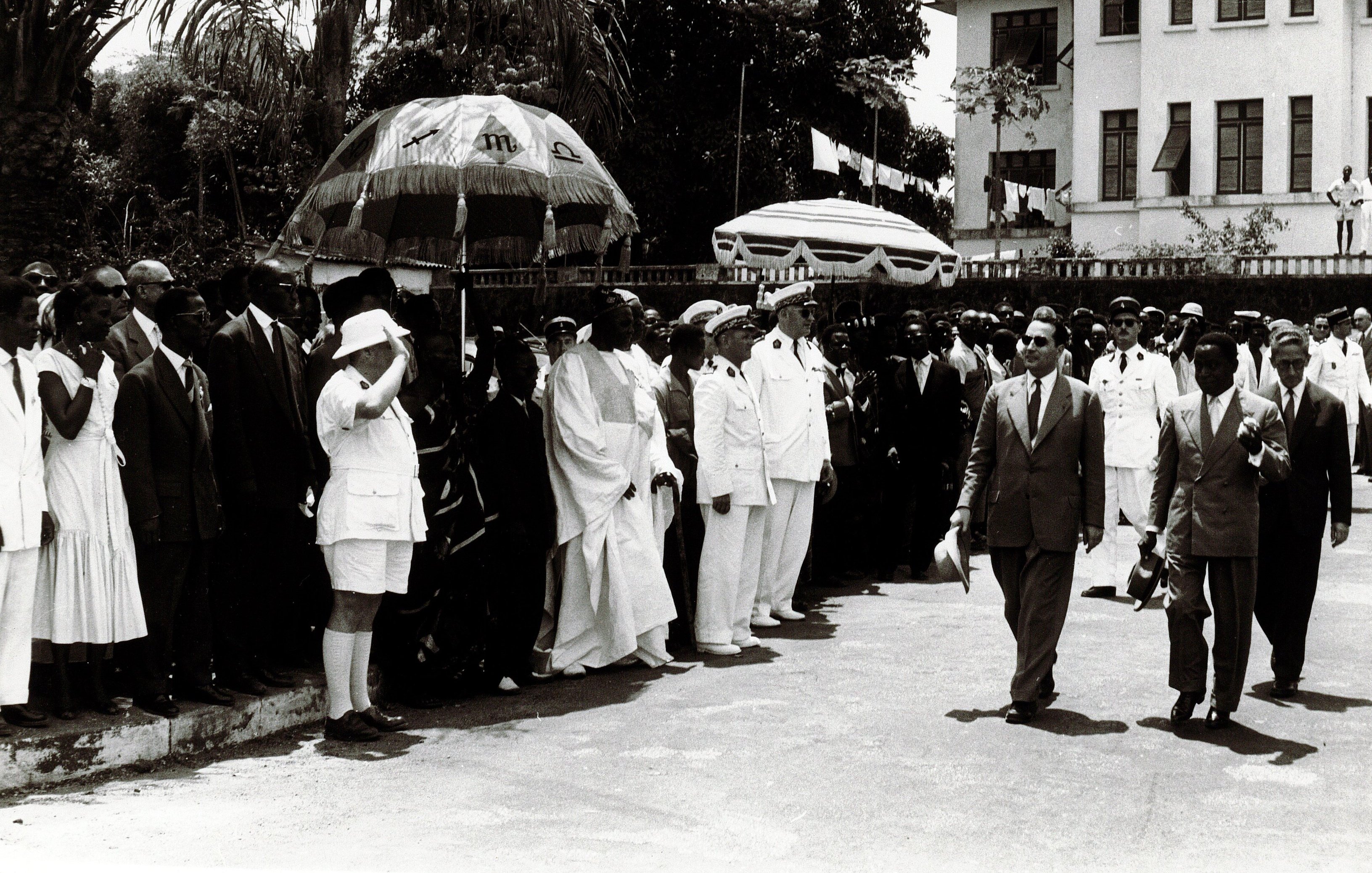 L'image montre un groupe de personnes rassemblées en extérieur, probablement lors d'un événement officiel. Au centre, des hommes en costumes formels marchent, entourés de dignitaires et de membres du public. Certains portent des uniformes militaires, tandis que d'autres sont habillés de manière civile. Il y a également des parapluies décoratifs en arrière-plan. L'atmosphère semble solennelle, et l'environnement suggère une atmosphère tropicale avec des arbres en arrière-plan. La scène donne une impression de cérémonie et de célébration.