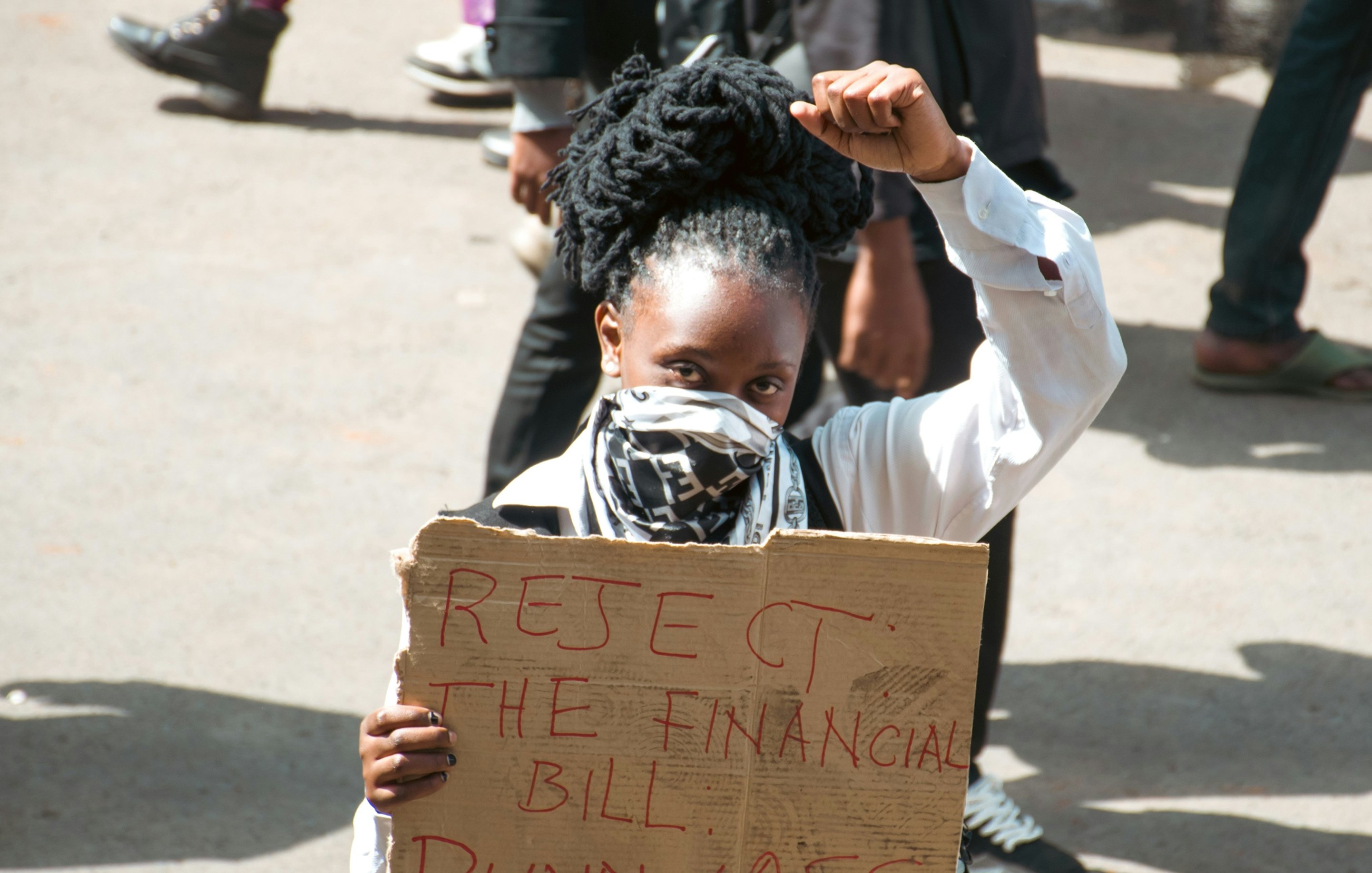 The image depicts a protester holding a cardboard sign that reads "REJECT THE FINANCIAL BILL PUNNYASS." The individual is wearing a white shirt and has their hair styled in large, coiled forms. They are raising a fist in a gesture of defiance or solidarity, and are also wearing a mask over their face. The background includes other people and a busy street scene, suggesting a gathering or demonstration.