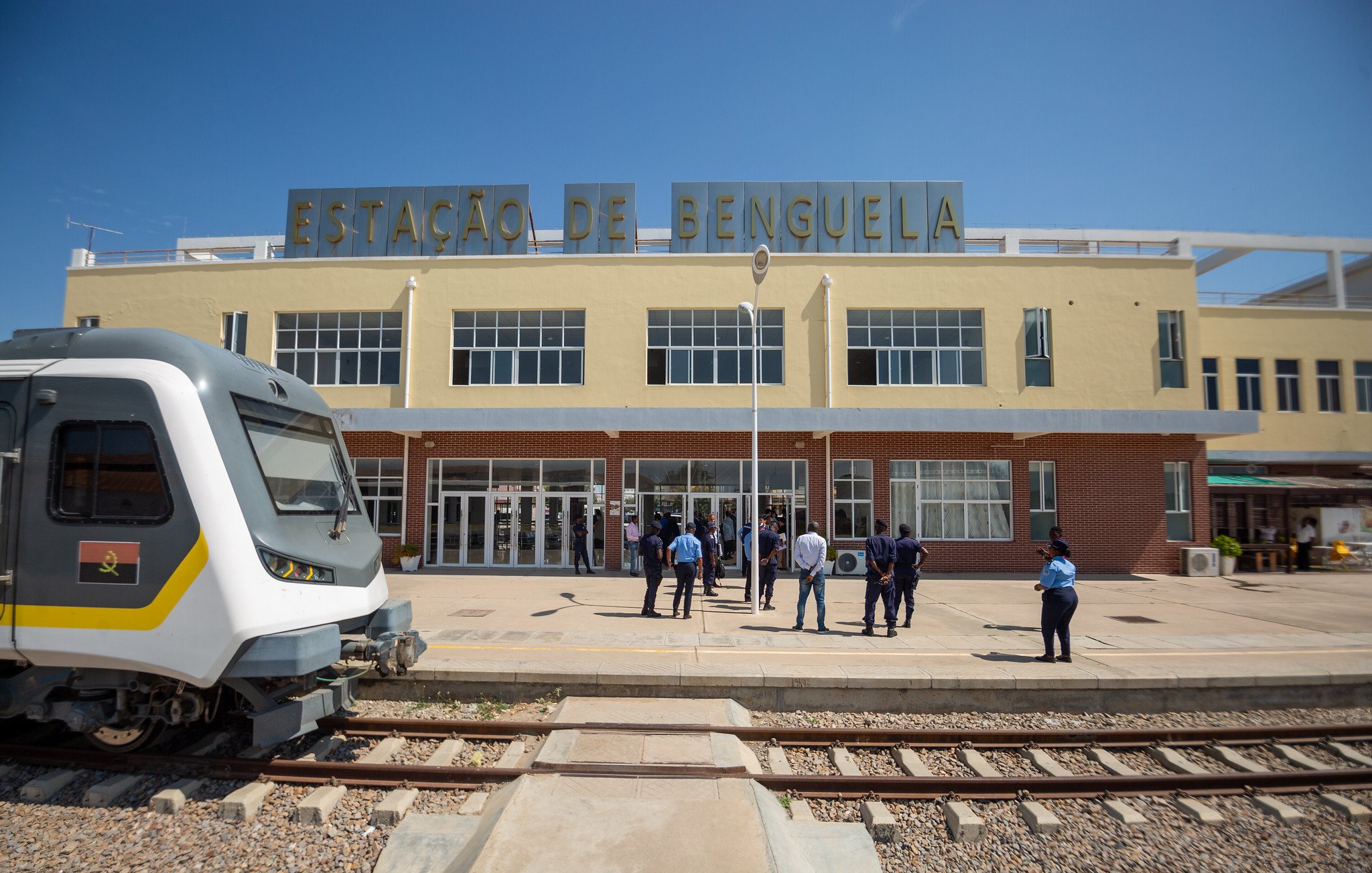 The image shows the exterior of a train station labeled "ESTAÇÃO DE BENGUELA." The building has a modern architectural design with large glass windows and a clean facade. In front of the station, a group of people, likely staff or passengers, are gathered. To the left, there is a sleek train parked on the tracks, showcasing a contemporary design. The scene is bright, indicating a clear blue sky. The overall setting suggests a bustling transportation hub.