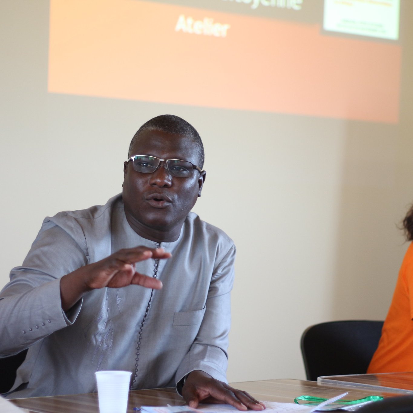 L'image représente un atelier se déroulant dans une salle de réunion. Au premier plan, deux personnes sont assises à une table. À gauche, un homme portant une chemise traditionnelle grise. Il semble s'exprimer avec enthousiasme, levant une main pour souligner un point important. À droite, une femme avec des cheveux bouclés est assise, portant un t-shirt orange. Elle l'écoute attentivement. En arrière-plan, un écran présente le mot "Atelier" en lettres d'origine orange. La scène est empreinte d'une atmosphère collaborative et interactive.