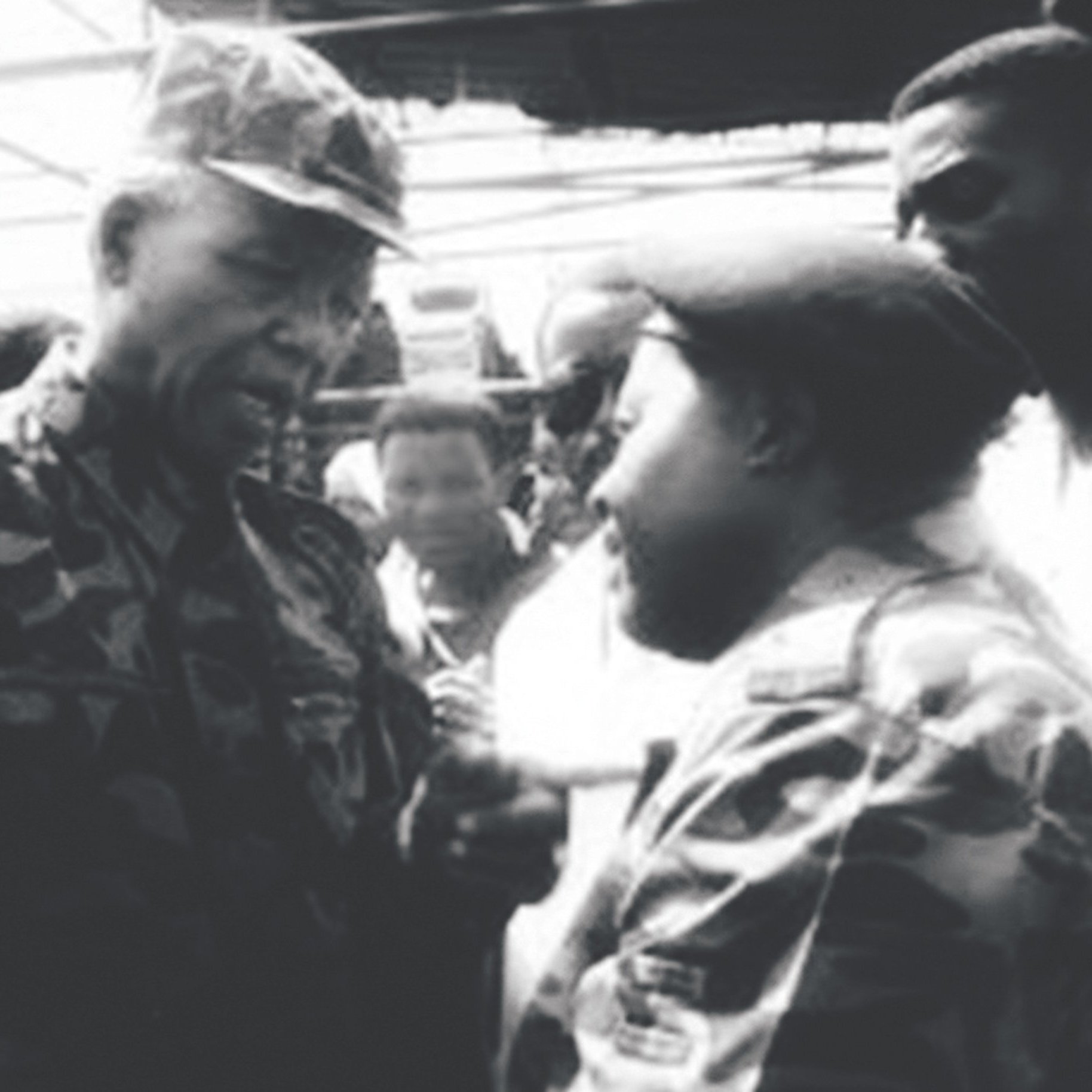 The image appears to be a historic black-and-white photograph depicting two men engaged in a conversation. One man is wearing military camouflage and appears to be in a position of authority, while the other is also dressed in military attire. They are surrounded by a group of people, indicating a significant setting, possibly a public event or gathering. The expressions on their faces suggest a serious discussion. The context hints at a moment of political or military importance, likely related to South Africa's history.
