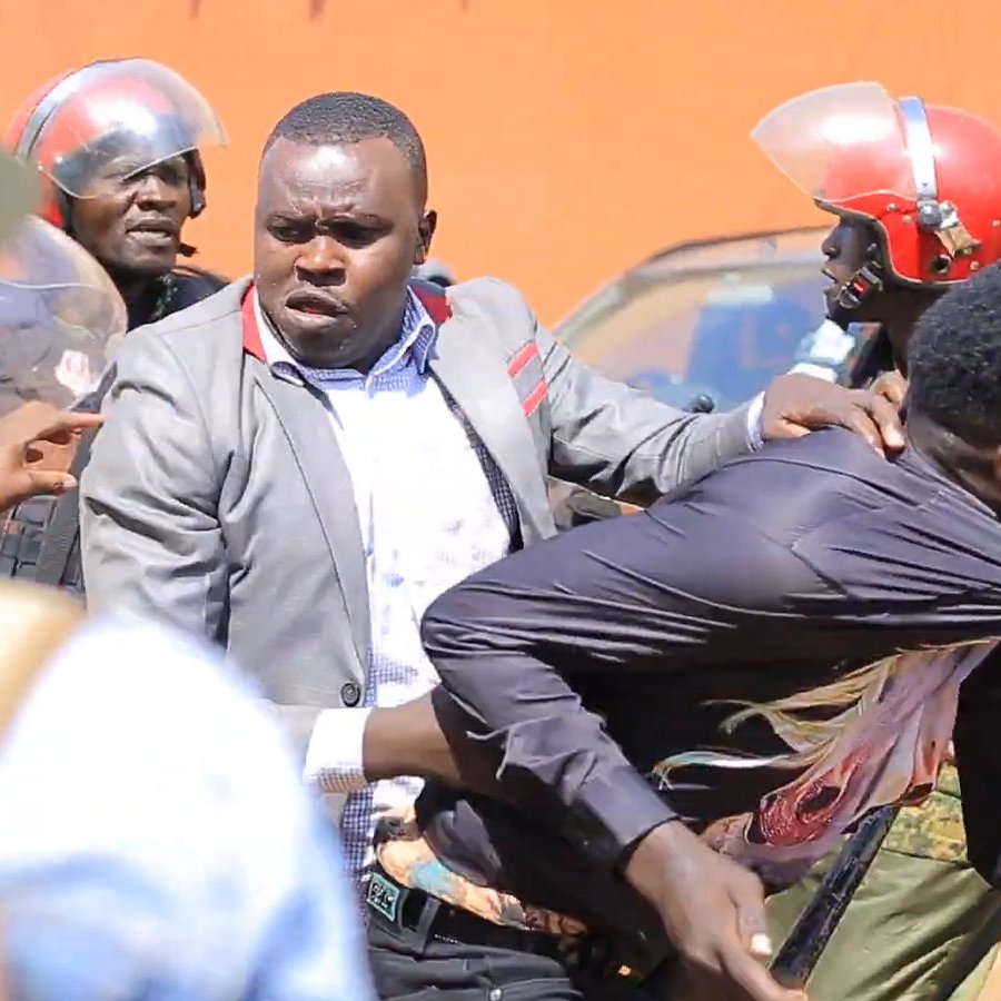 L'image montre une scène tendue sur une rue. On peut voir des policiers en uniforme, portant des casques rouges, qui semblent intervenir auprès d'un homme. Ce dernier est tiré en arrière par un autre homme qui essaie de le protéger. L'atmosphère est chaotique, avec des tensions visibles entre les forces de l'ordre et la personne qu'ils maîtrisent. En arrière-plan, il y a quelques personnes observant la situation et des véhicules garés. Le paysage évoque une ambiance de manifestation ou d'émeute.
